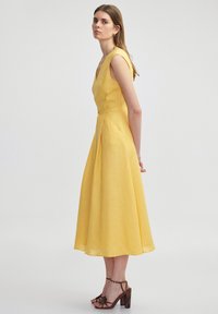 Żółta lniana sukienka z dekoltem w kształcie litery V, bez rękawów i plisowanym dołem, w zestawieniu z brązowymi sandałami na obcasie. Gładka faktura i długość do kolan.