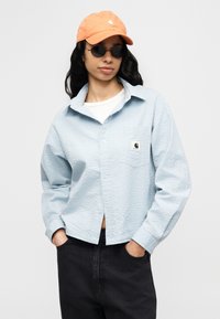 Ανοιχτό μπλε πουκάμισο με υφή και κουμπιά, με τσέπη στο στήθος και μικρό λογότυπο, συνδυασμένο με λευκό t-shirt και μαύρο παντελόνι. Περιλαμβάνει πορτοκαλί καπέλο και γυαλιά ηλίου.