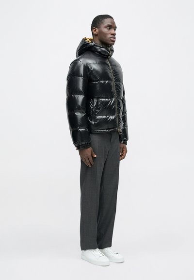 Dunjakke i skinnende sort materiale med hætte, med en gulddragelås og mønstret indvendigt for. Båret med grå bukser og hvide sneakers.