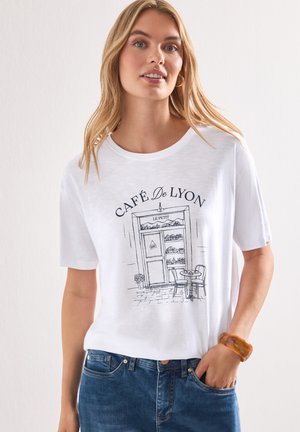 Femme blonde portant un t-shirt blanc avec un croquis noir de la devanture d'un café et "CAFÉ De LYON" imprimé au-dessus.
