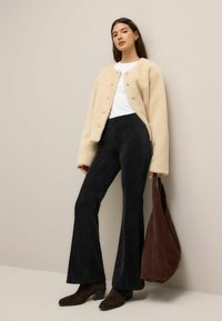 Beige Kunstfelljacke mit rundem Ausschnitt und Knopfverschluss, kombiniert mit schwarzen Schlaghosen, braunen Wildlederstiefeln und einer braunen Tragetasche.