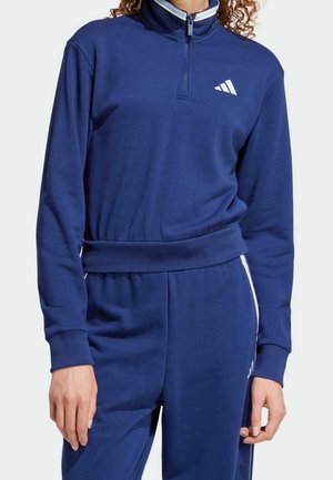 Osoba ubrana w granatowy sportowy dres z krótkim zamkiem i białym logo na piersi oraz spodniach.