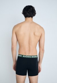 Fekete boxer alsónadrág zöld derékrésszel, rajta a "BJÖRN BORG" felirattal. Az anyag simának tűnik, testhezálló fazonnal és combközép hosszúsággal. Hátulról nézve.