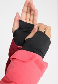 Mains portant des gants noirs sans doigts avec des ouvertures pour le pouce, ajustant les poignets d'une manche de veste rouge et rose.