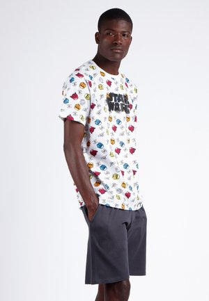 Camiseta blanca con estampados de dibujos animados coloridos, que presenta un logo negro de "STAR WARS". Combinada con pantalones cortos gris oscuro. El modelo está en pose con las manos en los bolsillos.