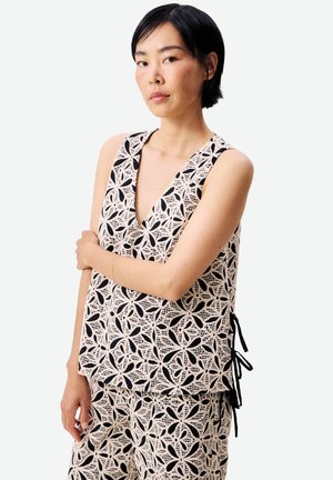 Femme aux cheveux courts noirs portant un haut sans manches en dentelle beige et un pantalon assorti avec des motifs floraux noirs, debout les bras croisés.