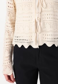 Pletený cardigan v off-white barvě s otvorenými krajkovými vzory, vlnkovaným lemem a stahovací šňůrkou. V kombinaci s černými kalhotami.