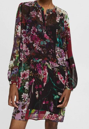 Femme portant une robe à manches longues, boutonnée sur le devant, à motif floral avec un mélange de couleurs sombres et vives et un tissu transparent.