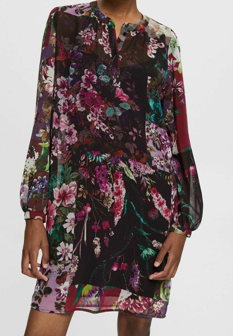 Femme portant une robe à manches longues, boutonnée sur le devant, à motif floral avec un mélange de couleurs sombres et vives et un tissu transparent.