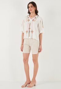 Top blanco con acentos bordados coloridos, combinado con pantalones cortos de lino beige. La modelo lleva sandalias de tira transparente. Fondo minimalista.