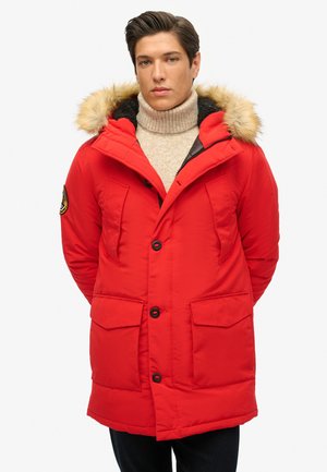 Parka isolée rouge avec une capuche bordée de fourrure, deux poches avant, fermeture à boutons et un col texturé. Présente des accents en métal noir.