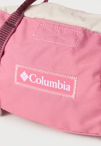 Ροζ υφασμάτινη τσάντα με υφή επιφάνειας, διαθέτει ραμμένη λευκή ετικέτα λογότυπου και αντίθετο κρεμ χρώμα στο πάνω μέρος. Ρυθμιζόμενο λουράκι.