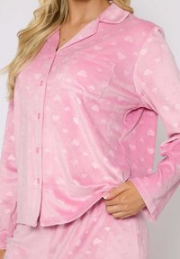 Conjunto de pijama rosa hecho de una tela suave con pequeños patrones de corazones blancos, que incluye una camisa con botones y un bolsillo en el pecho, además de una cintura elástica.