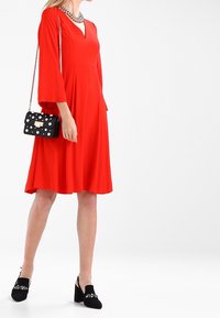 Abito rosso a-line con maniche a campana, scollatura a V e orlo svasato. Borsa nera con pois bianchi. Scarpe nere con tacco a blocco e decorazioni.