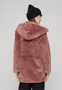 Persona, kas valkā putekļaini rozā mākslīgās kažokādas mēteli ar kapuci un melnu adītu cepuri, skatīta no muguras pret vienkrāsainu fonu.