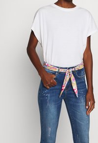 Bílý top s krátkým rukávem spárovaný s modrými džíny, které mají barevný vzorovaný pásek a dekorativní cvočky na kapsách.