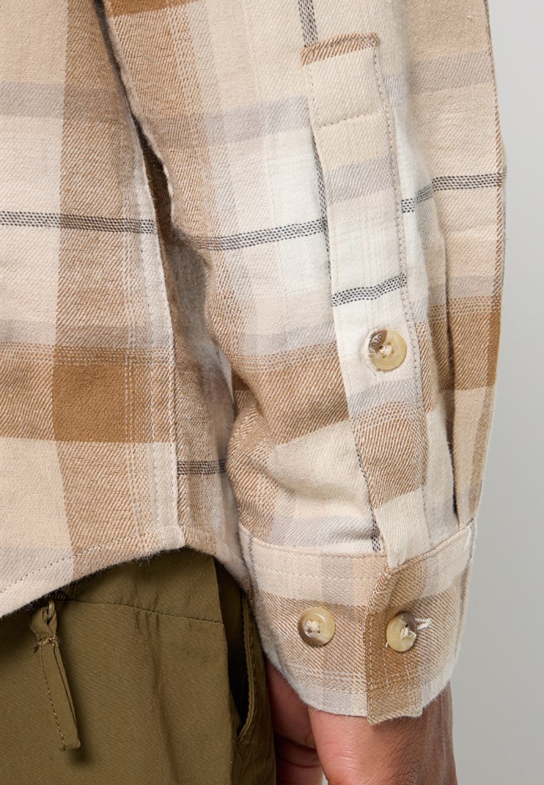 Manga de una camisa de cuadros en tono beige y marrón con dos botones en el puño, combinada con pantalones verde oliva que tienen un cierre y un bolsillo visibles.