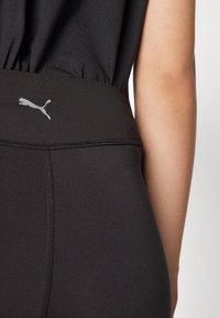 Crne sportske tajice s glatkom teksturom, s Puma logom u sivoj boji na pojasu i kontrastnim šavovima duž šavova.