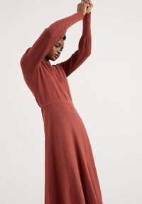 Abito a maniche lunghe di colore ruggine in materiale a maglia testurizzata, con un corpetto aderente e una gonna fluente, caratterizzato da dettagli a costine nelle maniche.