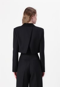 Crna skraćena blazer jakna s dugim rukavima, oštrim ovratnikom i jedinstvenim detaljem na leđima. Izrađena je od glatkog materijala s minimalističkim dizajnerskim elementima.