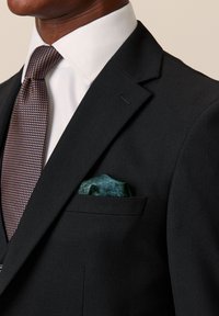 Abito nero con tessuto testurizzato, camicia bianca e cravatta marrone con motivo a fantasia. Fazzoletto da taschino con un motivo verde.