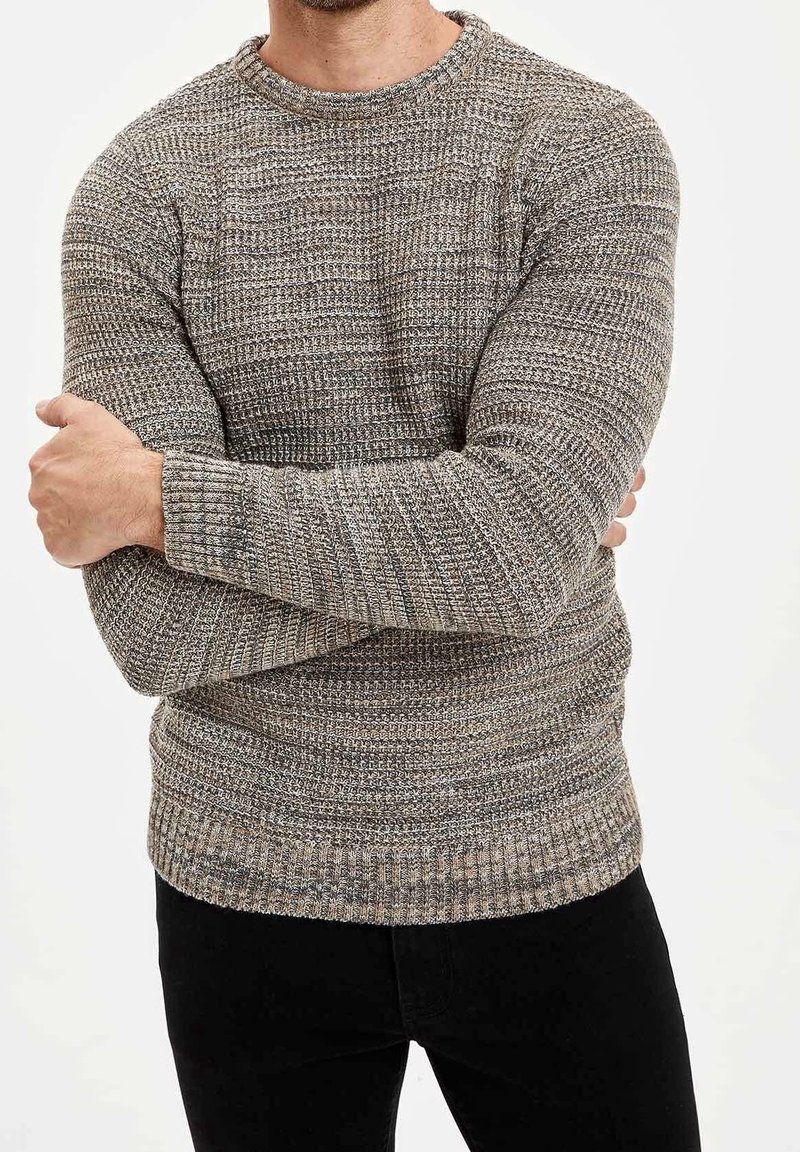 Homme portant un pull en maille texturé gris et marron à manches longues, bras croisés, associé à un pantalon noir.