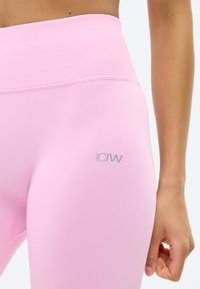 Κοντινή λήψη ενός ατόμου που φοράει ροζ κολάν με ψηλή μέση, με ένα μικρό αντανακλαστικό λογότυπο στο αριστερό μηρό και ένα εμφανές χέρι στο πλάι.