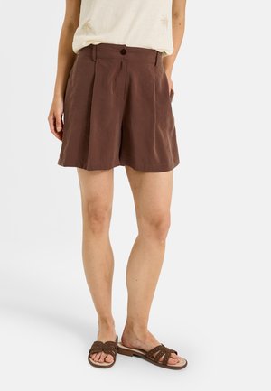 Femme portant un short marron taille haute avec des plis sur le devant, un haut beige sans manches et des sandales plates marron avec un design ajouré.