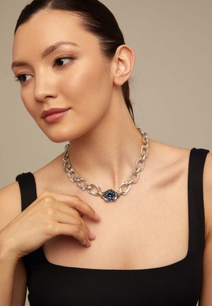 Mujer con cabello oscuro, vestida con una blusa negra sin mangas y un collar grueso de cadena plateada con una piedra preciosa azul en el centro.