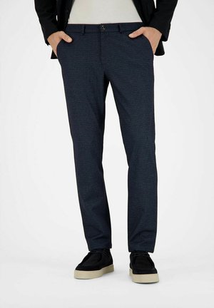 Man wearing slim dark blue textured trousers with hands in pockets and black shoes with white soles on white background.