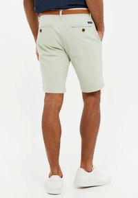 Pantalones cortos verde claro con una textura suave, dos bolsillos traseros y un cinturón trenzado marrón. Combinados con zapatillas blancas.