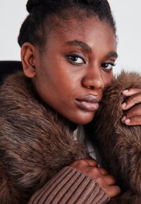 Λήψη κοντινού πλάνου ενός ατόμου με σκούρο δέρμα που φοράει ένα καφέ γούνινο μπουφάν και ένα ριπτό καφέ πουλόβερ, κρατώντας τρυφερά τον γιακά του μπουφάν.