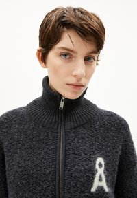 Pull à zip noir et texturé avec un col haut et une fermeture éclair argentée. Présente un logo "A" stylisé blanc sur la poitrine gauche.