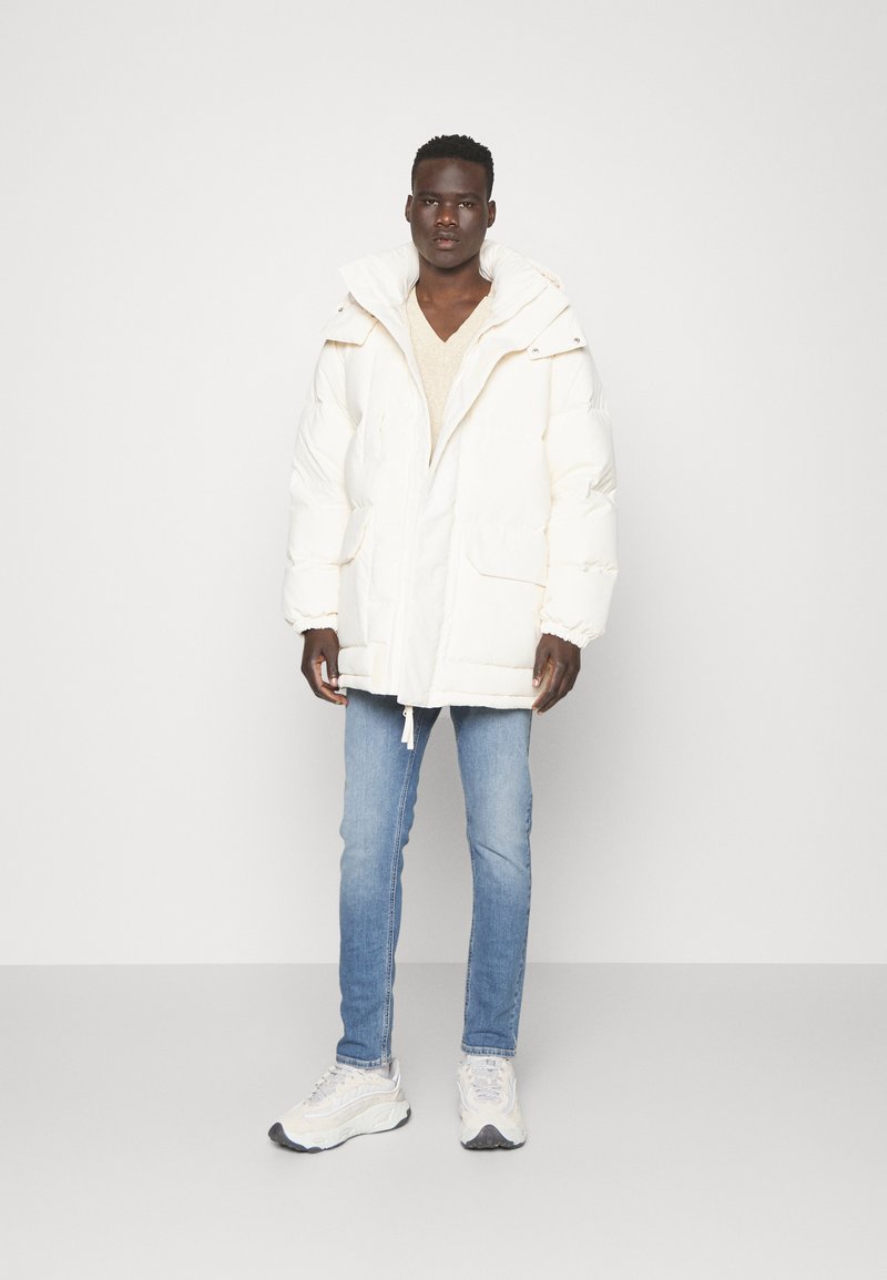 Parka blanche duveteuse avec une capuche, fermeture éclair à l'avant, deux grandes poches et poignets élastiques. Associée à un jean bleu clair et des baskets grises.