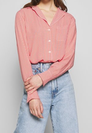 Mujer que lleva una blusa abotonada roja y blanca con lunares y mangas largas, metida en unos vaqueros azul claro de tiro alto.