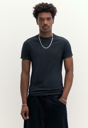 Giovane uomo con capelli ricci, indossa una semplice maglietta nera, una collana d'argento e braccialetti, in piedi con uno sfondo bianco.