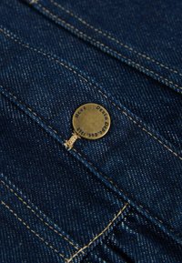 Tumši zila denima auduma, ar redzamu šuvju, kas satur misiņa pogu ar iegravētu tekstu un pievienotu plānu brūnu diegu.