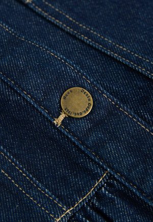 Dunkelblauer Denim-Stoff mit sichtbaren Nähten, ausgestattet mit einem Messingknopf mit eingraviertem Text und einem dünnen, hellbraunen Faden.