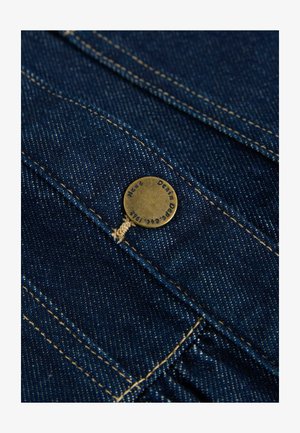 Tamsiai mėlynas džinsinis audinys su matomu siuvinėjimu, su bronzine pakabos sagtele, ant kurios yra iškalti užrašai, ir pritvirtinta plona tan spalvos siūle.