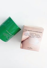 Zielony cylindryczny pojemnik z etykietą "Czysty Balsam do Grindingu" obok różowego pudełka z informacjami o składnikach, na którym z boku pokazana jest kremowa konsystencja.