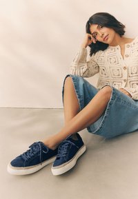Marineblå sneakers med overdel i tekstureret stof, hvid gummisål og kontrastfarvede snørebånd, kombineret med et lyst denimskørt og en strikket cardigan.