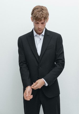 Schwarzer maßgeschneiderter Anzug mit Notch-Lapel, einem einzelnen Knopfverschluss und einem glatten weißen Hemd darunter. Justiert die Manschette der Jacke.