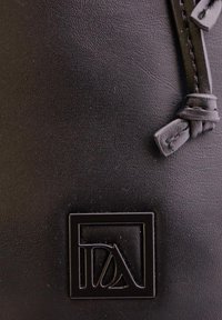 Superficie de cuero negro con una correa cosida atada en un nudo cerca de un logo cuadrado en relieve que presenta letras estilizadas en su interior.