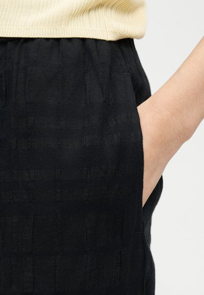 Χέρι που εδράζεται στην τσέπη μαύρων υφασμάτινων παντελονιών, φορώντας ανοιχτό μπεζ ριμπ μπλουζάκι, απεικονιζόμενο από τη μέση μέχρι το μέσο του μηρού πάνω σε ουδέτερο φόντο.