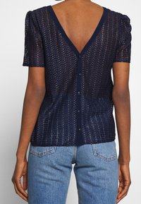 Haut bleu marine à manches courtes avec un décolleté en V, présentant un motif tricoté texturé et des boutons le long du dos.