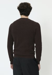Pull en maille marron foncé avec manches longues, col rond, et détails côtelés aux poignets et à l'ourlet, porté avec un pantalon noir.