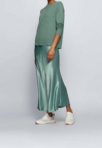 Grön tröja med lång ärm, parat med en glänsande, silkesliknande tealkjol och vita sneakers med beige detaljer. Släta texturer dominerar outfiten.