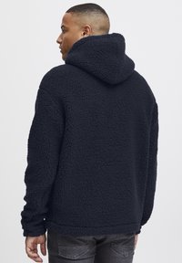 Marineblå fleece hettegenser med en teksturert, myk overflate. Har en stor hette og ribbestrikkede mansjetter. Fremstår varm og uformell i design.