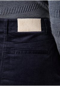 Pantaloni in velluto a coste di un blu scuro, con una fascia in vita strutturata e una toppa di tessuto color crema sul retro. Presentano una tasca posteriore con cuciture orizzontali.