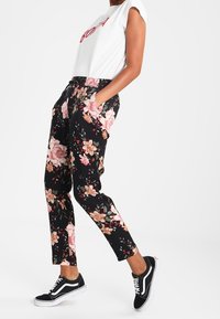 Person som bär svarta blommiga byxor, en vit ärmlös topp med röd text och svart-vita sneakers, står med händerna i fickorna.